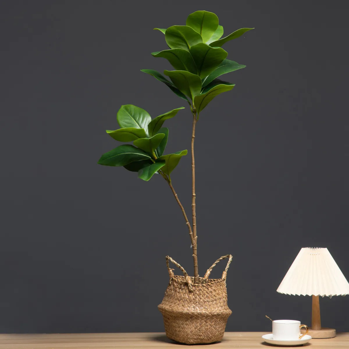 Faux Magnolia Tree Branch in woven basket on a wooden surface with a lamp and cup.