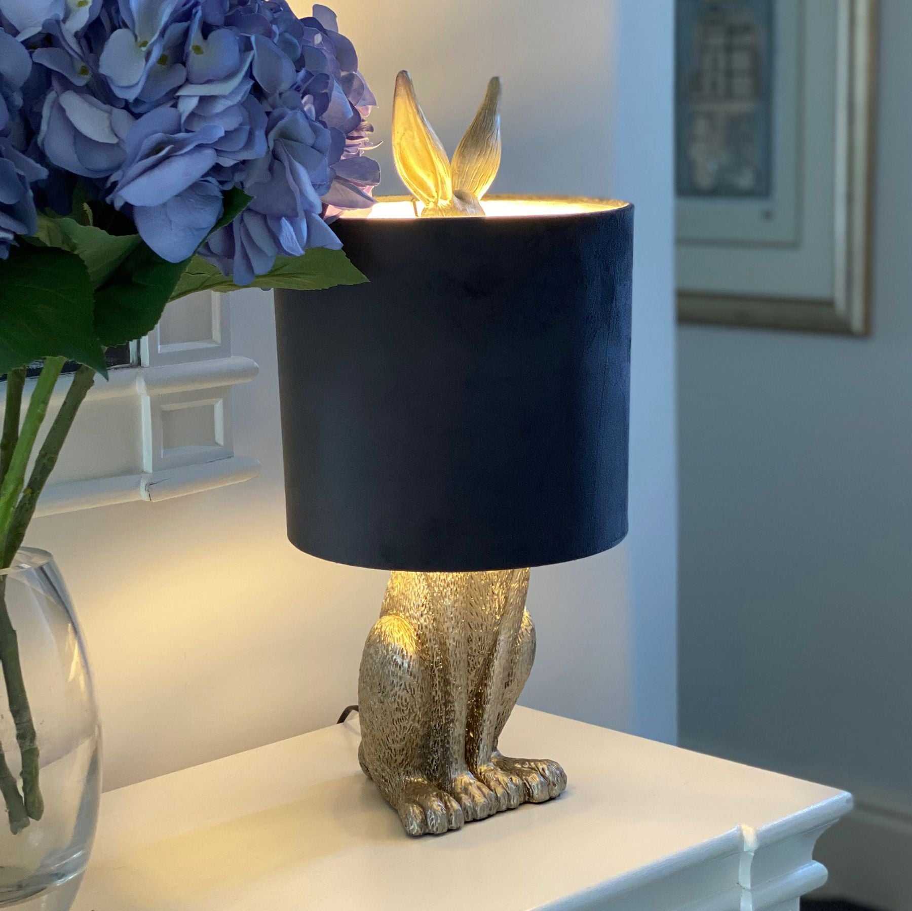 Silver rabbit table lamp with grey velvet shade on a white surface.